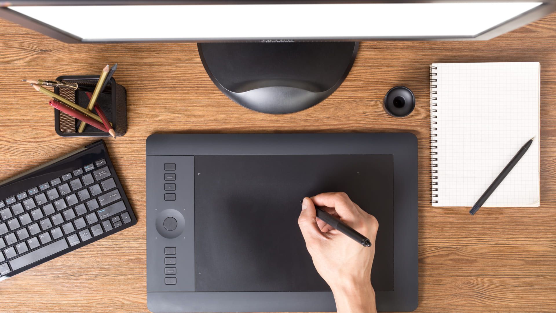 Mesa digitalizadora preta com teclado sobre mesa de madeira com outros acessórios de trabalho próximos