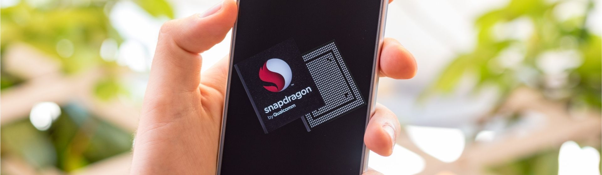 Celular com a logo do Snapdragon na mão de homem com plantas ao fundo