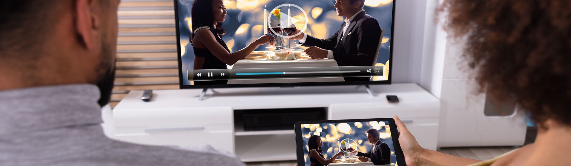 Casal negro de costas sentado em frente a uma TV e segurando tablet, com conteúdo espelhado para a tela da TV.