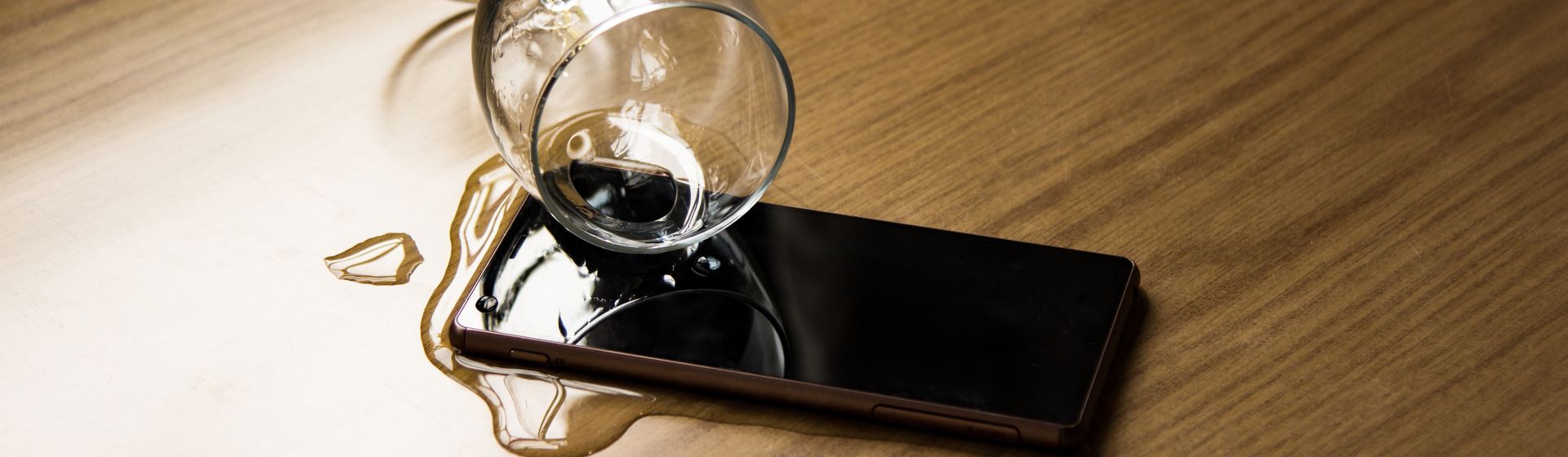 Copo de vidro entornando água em um celular em cima de uma mesa de madeira