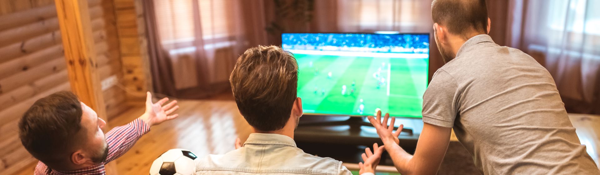 Três homens brancos em frente a uma TV assistindo a um jogo de futebol e torcendo.