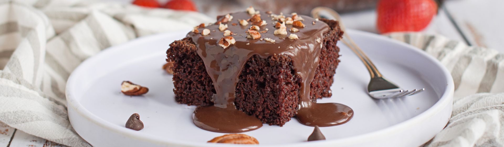 Pedaço de bolo de chocolate de liquidificador com calda de chocolate e farofa de castanhas, em um prato branco com garfo.