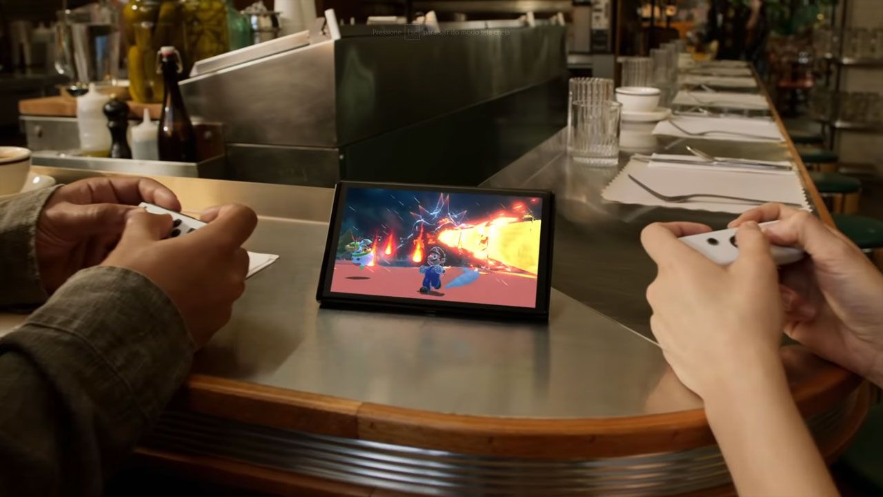 Nintendo Switch OLED no centro, com jogo na tela e duas mãos segurando os controles enquanto jogam.