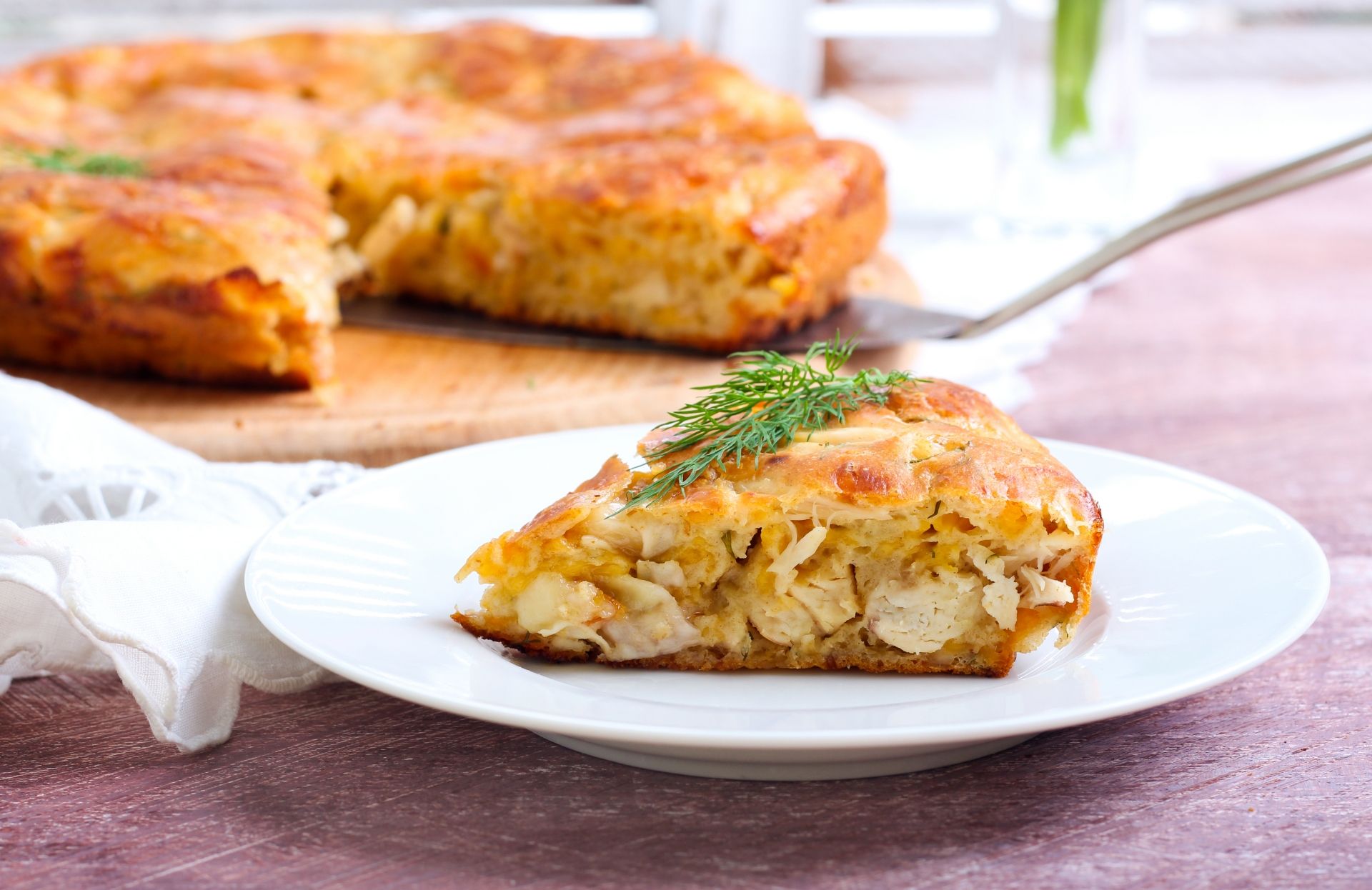 Fatia triangular de torta de frango num prato com a torta inteira ao fundo