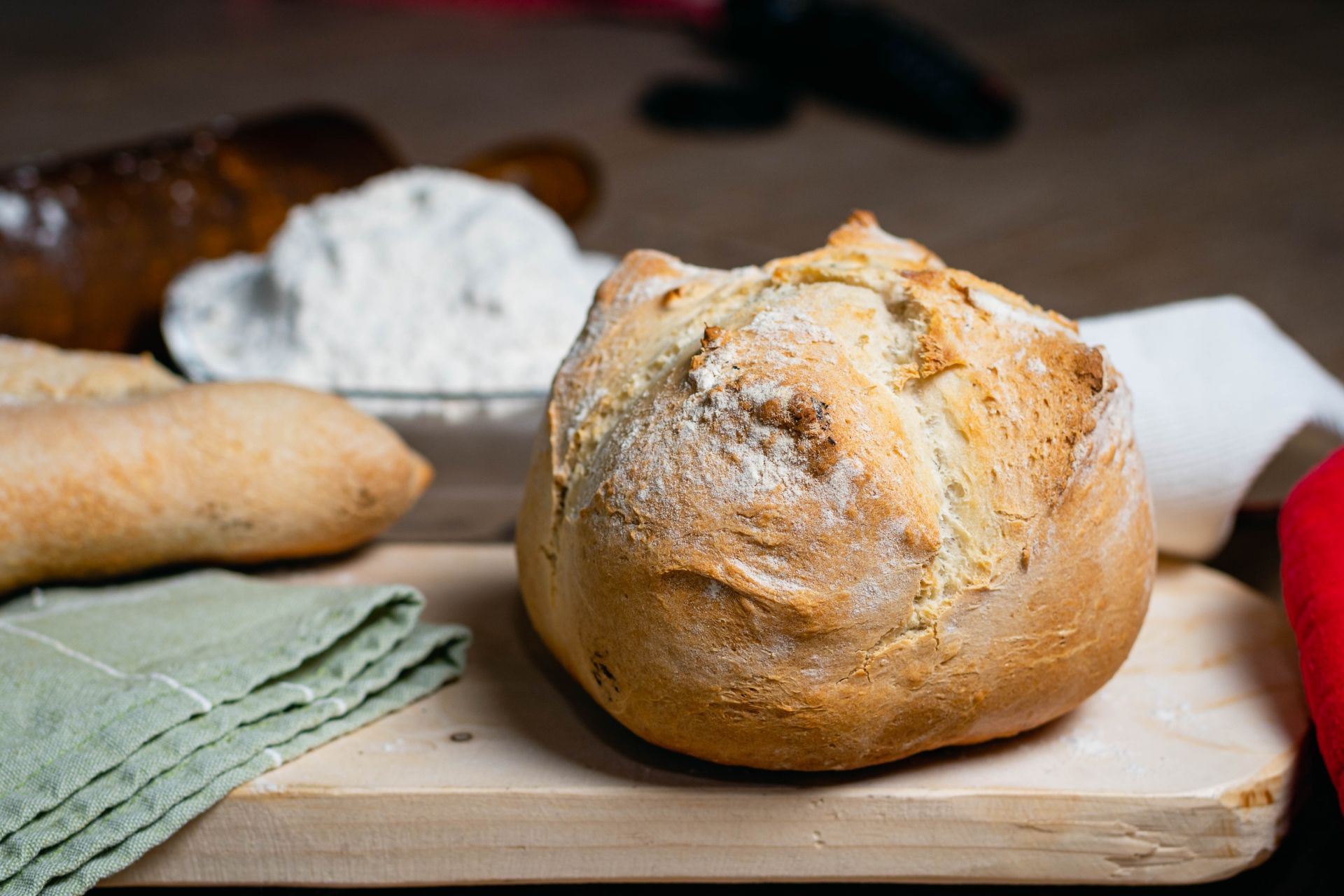 Pão caseiro redondo numa tábua com ingredientes na frente de um recipiente com farinha de trigo