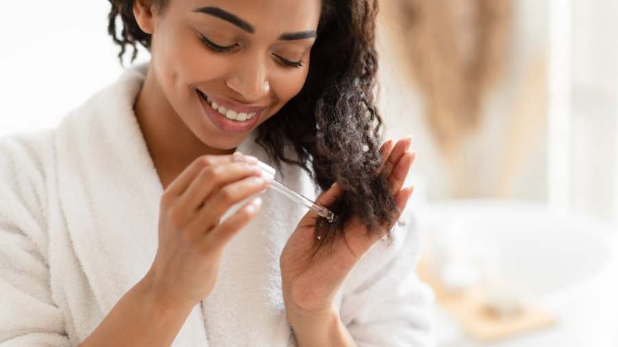 Mulher de roupão branco sorrindo enquanto aplica óleo para cabelo nos fios