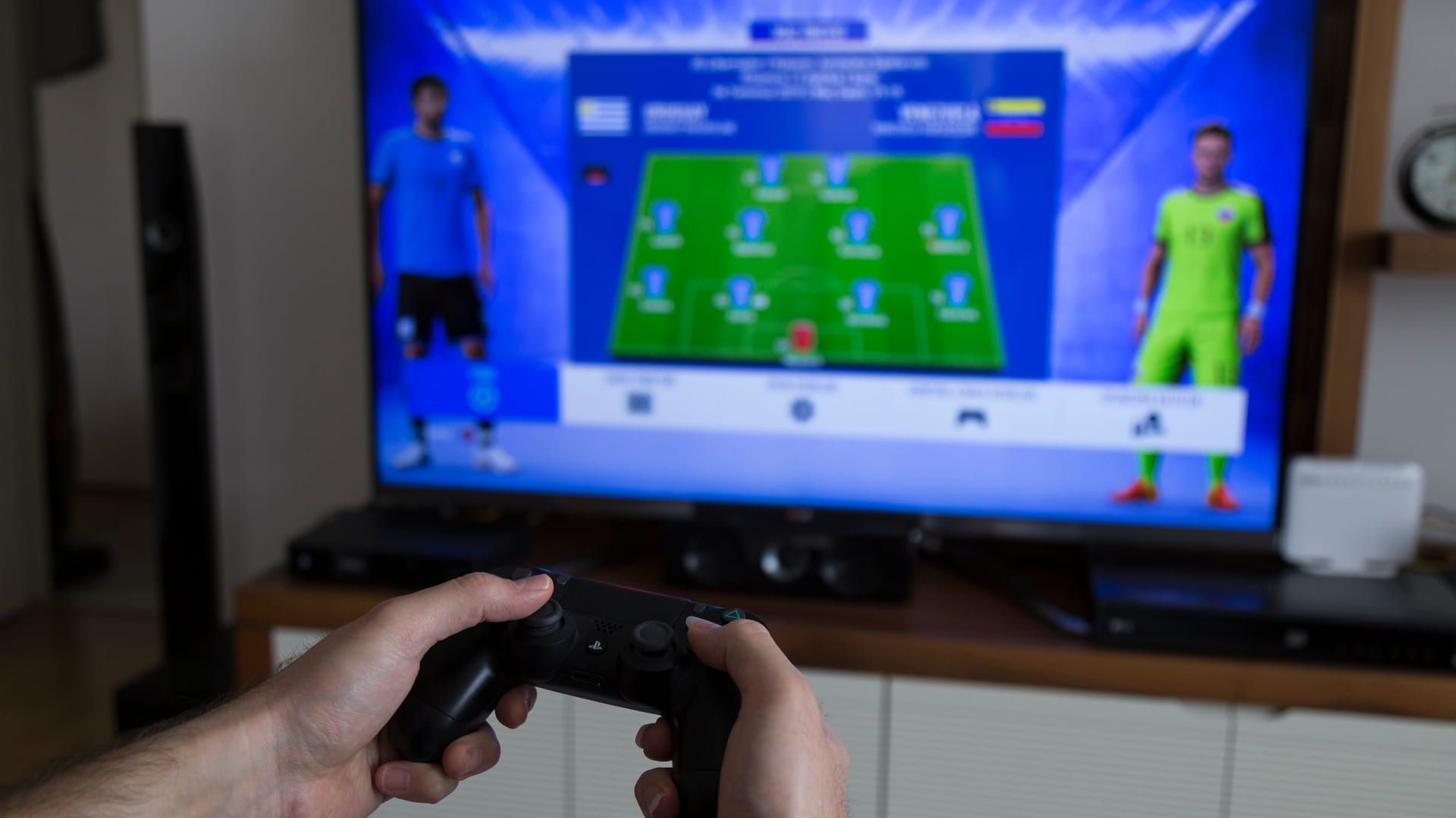 Homem jogando com controle de PS4 com jogo de futebol sendo exibido em TV no fundo desfocado da tela