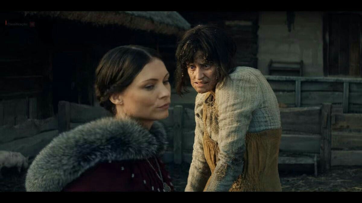 Yennefer de Vengerberg, no plano de fundo e com suas deficiências, usando roupa de camponesa, enquanto no primeiro plano está Tissaia de Vries, reitora da sua escola de magia, com roupas chiques