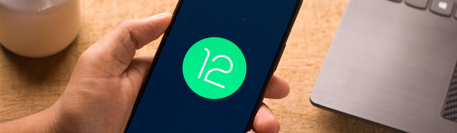 Celular em mão de homem com Android 12 na tela, notebook e item de decoração ao fundo