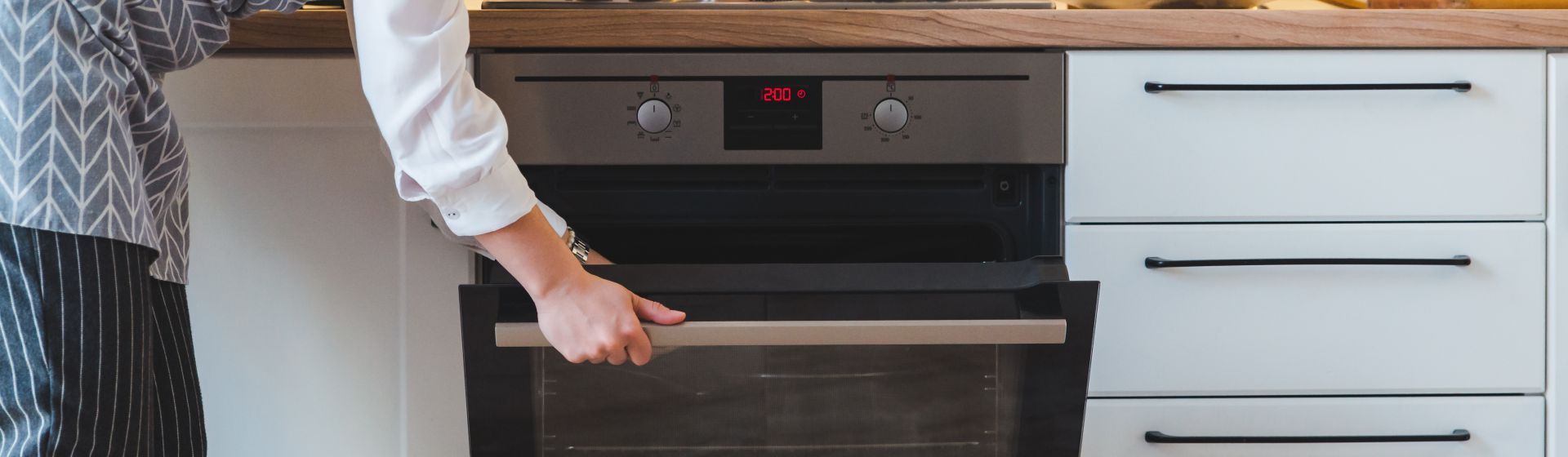 Mulher abrindo forno inox embutido na bancada da cozinha. Forno fica entre armários e gavetas brancas e tampo de madeira