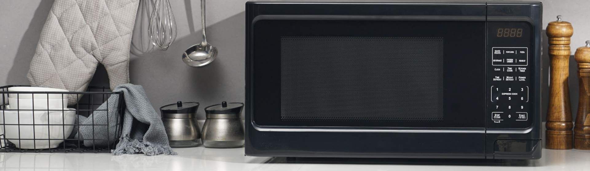 Micro-ondas preto em cima de bancada branca e entre utensílios de cozinha diversos