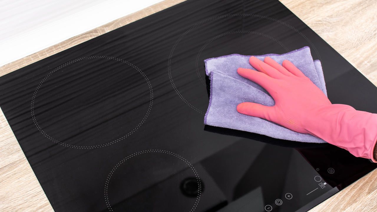 Mão com luva cor de rosa segurando um paninho lilás enquanto limpa um fogão de indução preto