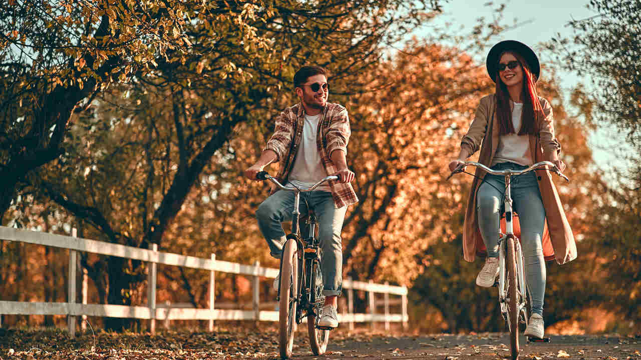 Homem e mulher jovens usando jeans, camiseta branca e blusa marrom, andando de bicicleta lado-a-lado em parque no outono