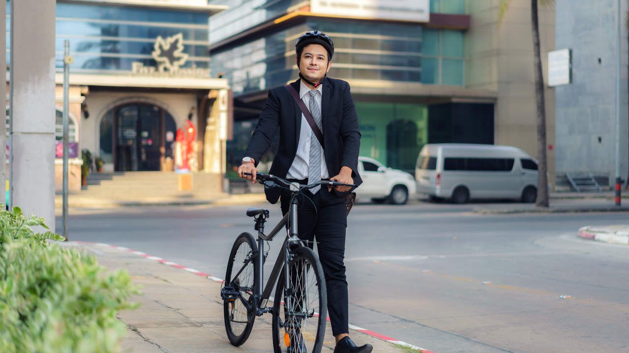 Homem asiático usando terno e capacete para ciclismo, carregando sua bicicleta lateralmente na calçada de um centro urbano, preparando-se para ir ao trabalho de manhã