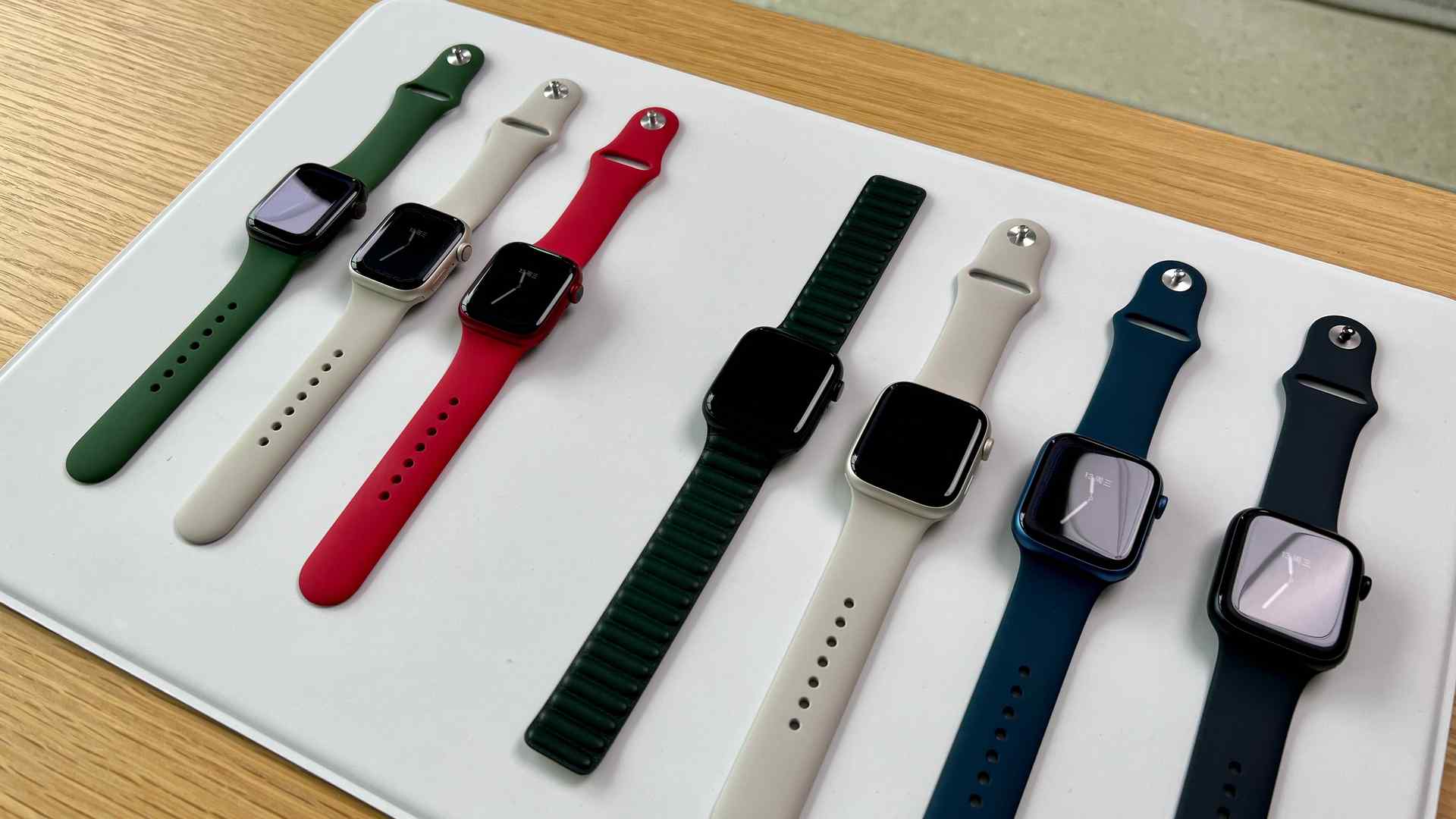 Sete opções de Apple Watch Series 7 de diferentes cores em suporte branco em mesa de madeira