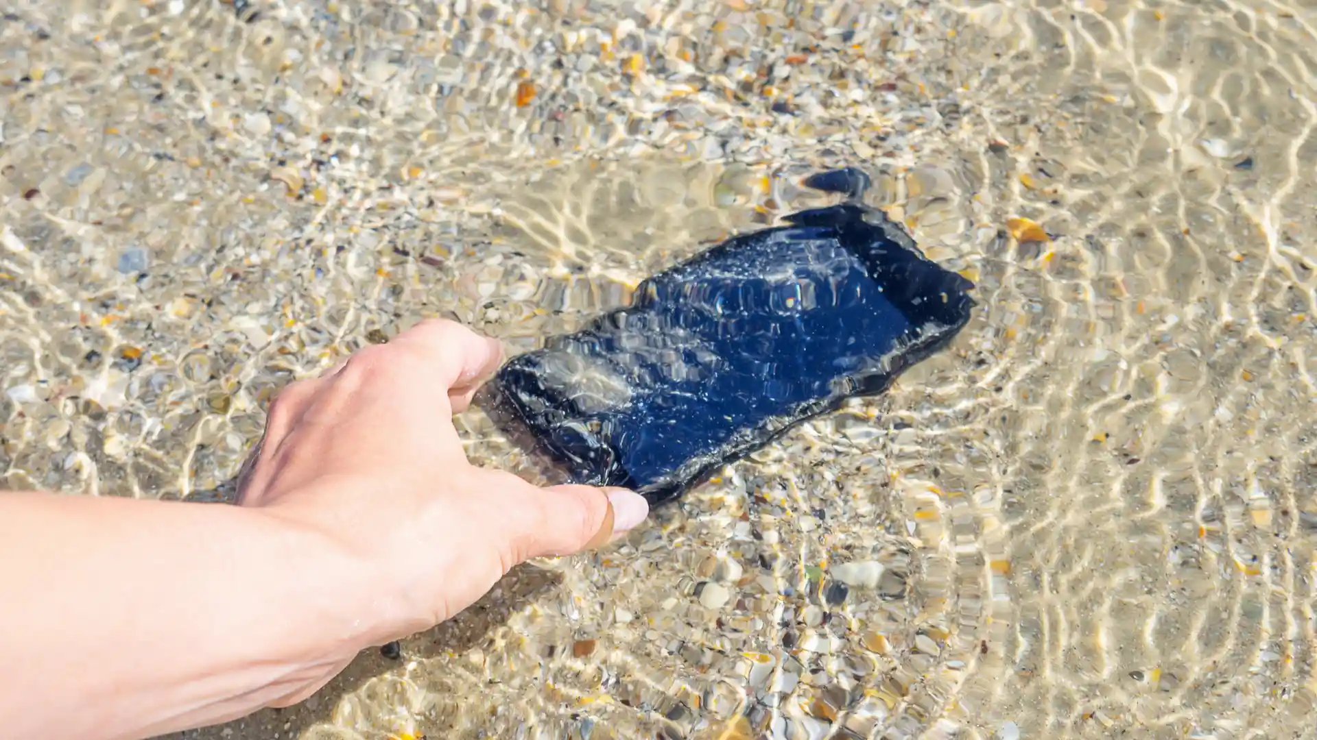 Pessoa pegando um celular submerso na água com areia e pedras ao fundo de dia