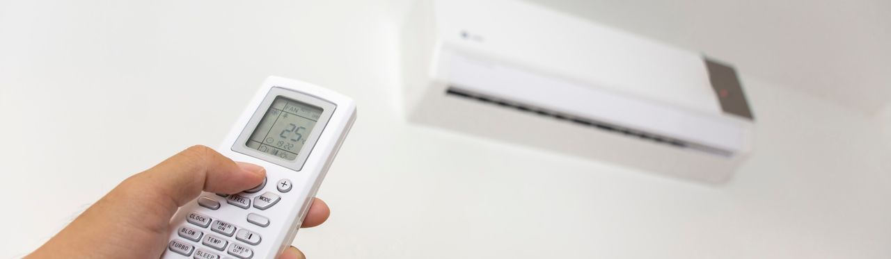 Em primeiro plano, uma mão apontando um controle remoto para um ar-condicionado, desfocado em segundo plano