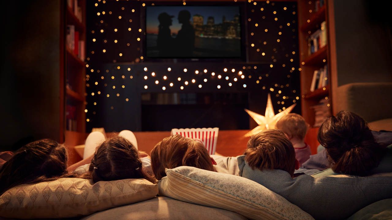 Pessoas deitadas com a cabeça no travesseiro em sala escura com TV, balde de pipoca e lâmpadas de LED espalhadas 