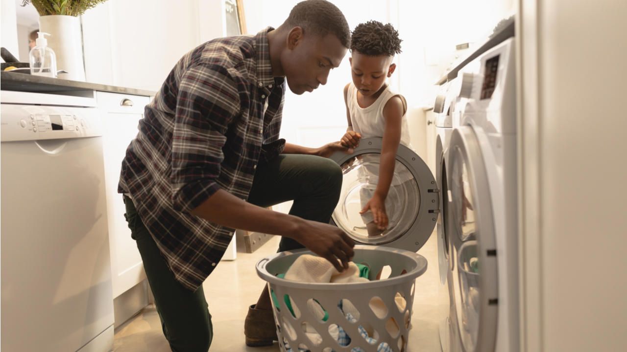 Pai e filho negros em lavanderia com cesto de roupa suja, colocando as peças em máquina de lavar front load 
