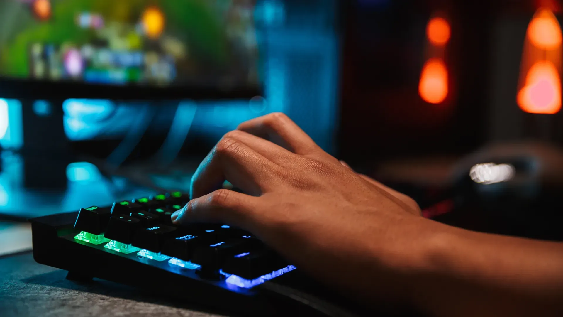 Um teclado preto em um ambiente pouco iluminado, onde podemos ver uma das mãos de uma pessoa branca teclando no aparelho, desfocada ao fundo e à esquerda está uma tela com o que parece ser o jogo League of Legends