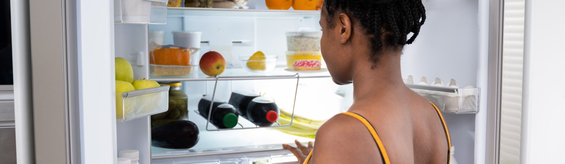 Mulher negra mexendo em uma geladeira de duas portas verticais com bebidas e frutas