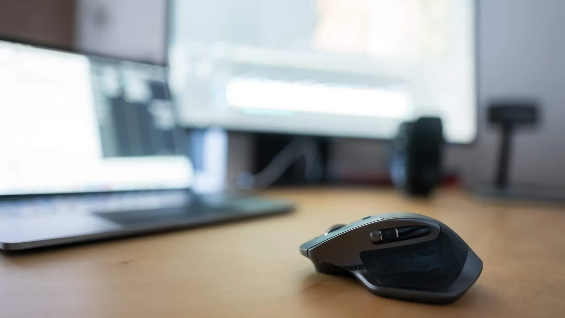 Mouse gamer cinza sobre mesa de madeira, com notebook e monitor desfocado ao fundo