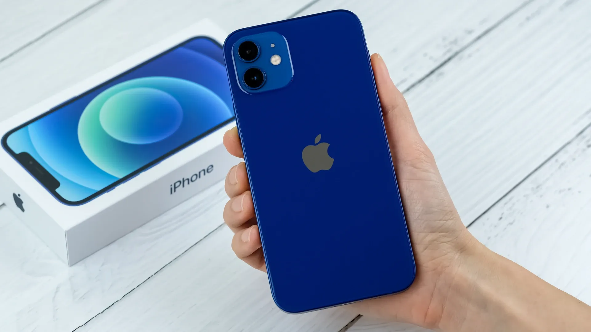 Mão segurando o iPhone 12 azul com caixa ao fundo