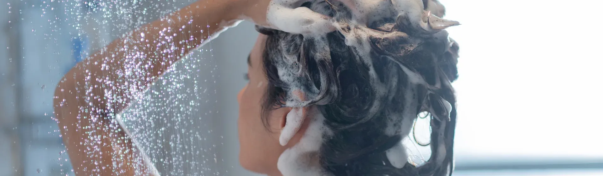 Mulher no banho, aplica shampoo no cabelo já com espuma, com o chuveiro ligado.