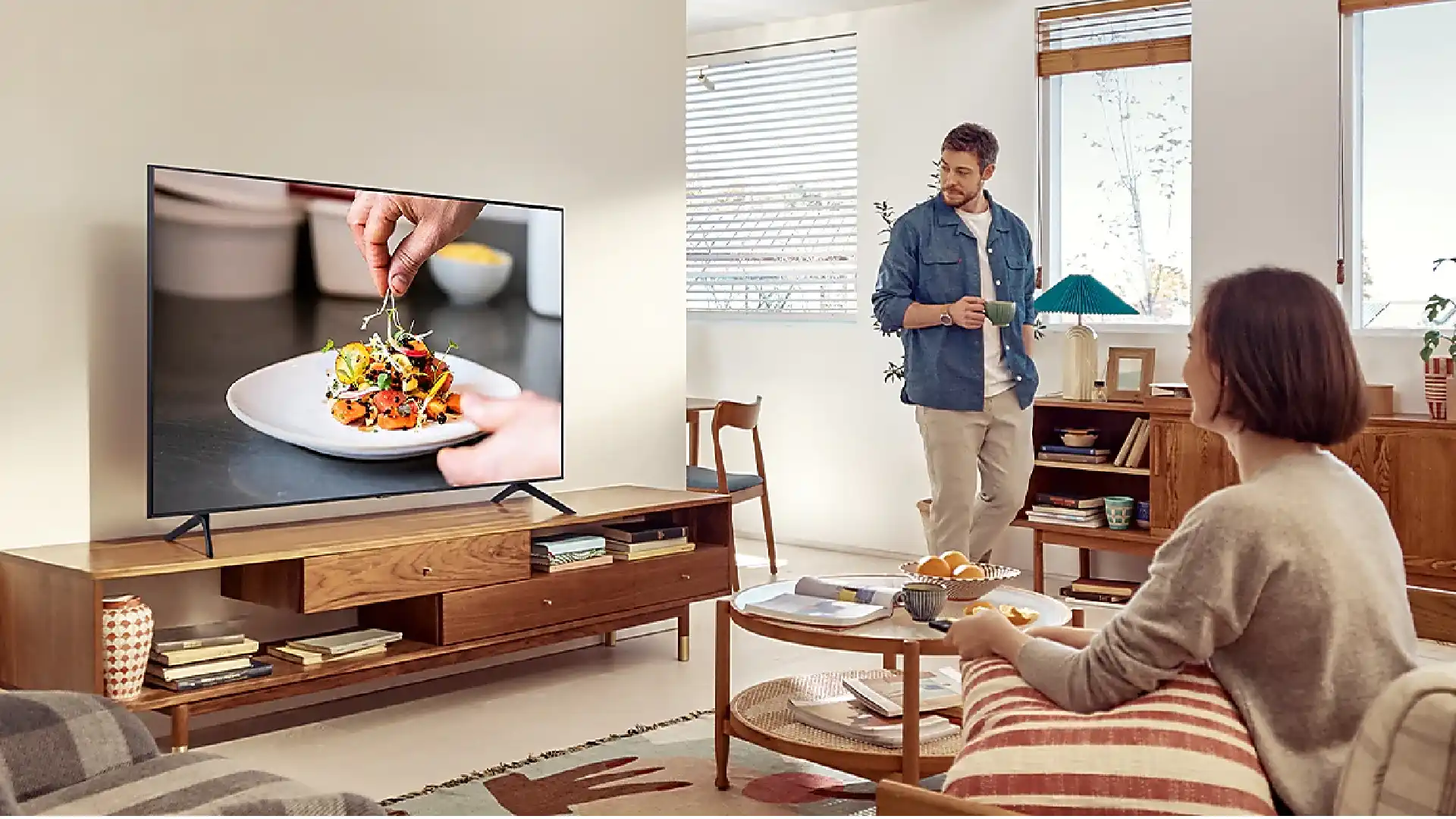 TV Samsung AU7000 sobre rack de madeira em sala de estar. Homem e mulher assistem à TV.