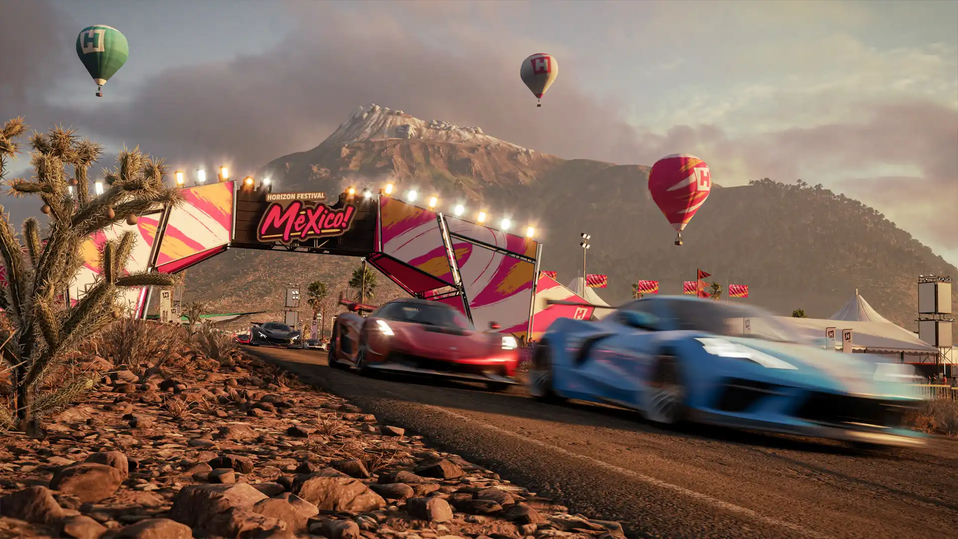 Imagem do jogo Forza Horizon 5 com um carro azul embaçado pela velocidade, um carro vermelho atrás dele e um terceiro carro preto mais atrás competindo em uma estrada de terra com montanhas e balões ao fundo e um letreiro escrito "Mexico!"
