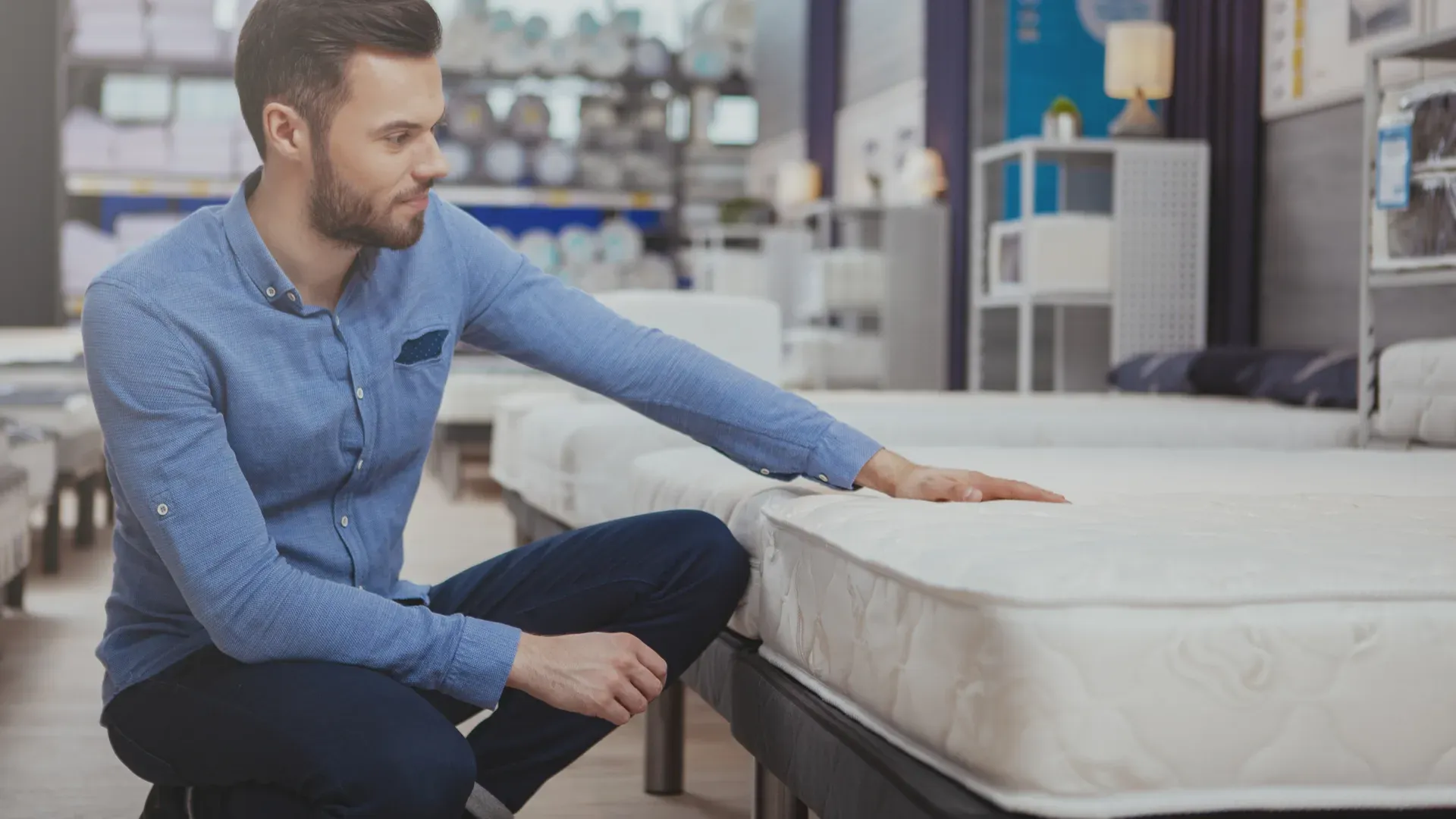 Homem agachado ao lado de cama em loja de móveis, com a mão sobre um colchão de solteiro