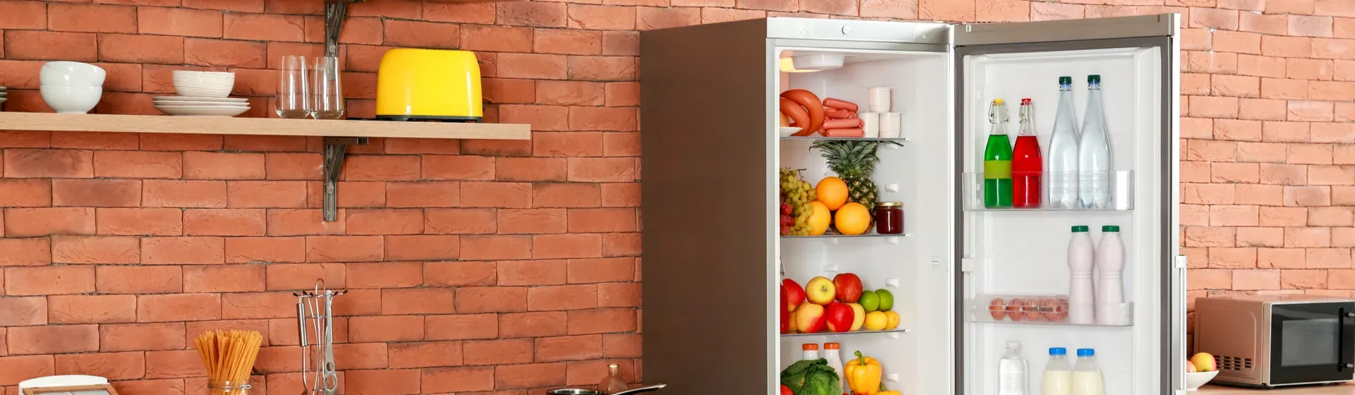 Cozinha com parede de tijolos e geladeira barata inox com a porta aberta e legumes e frutas no interior