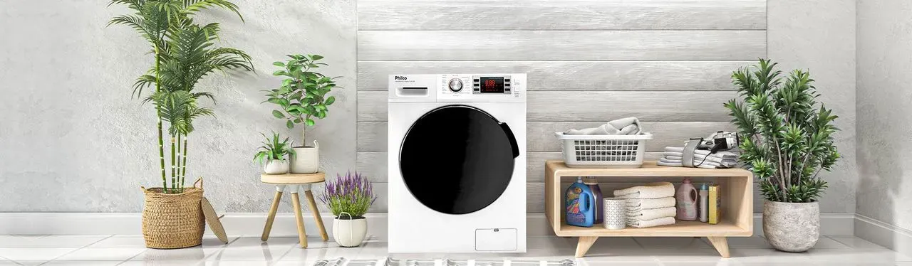 Máquina de lavar branca com abertura frontal e porta de vidro preto, em área de serviço de parece cinza com plantas