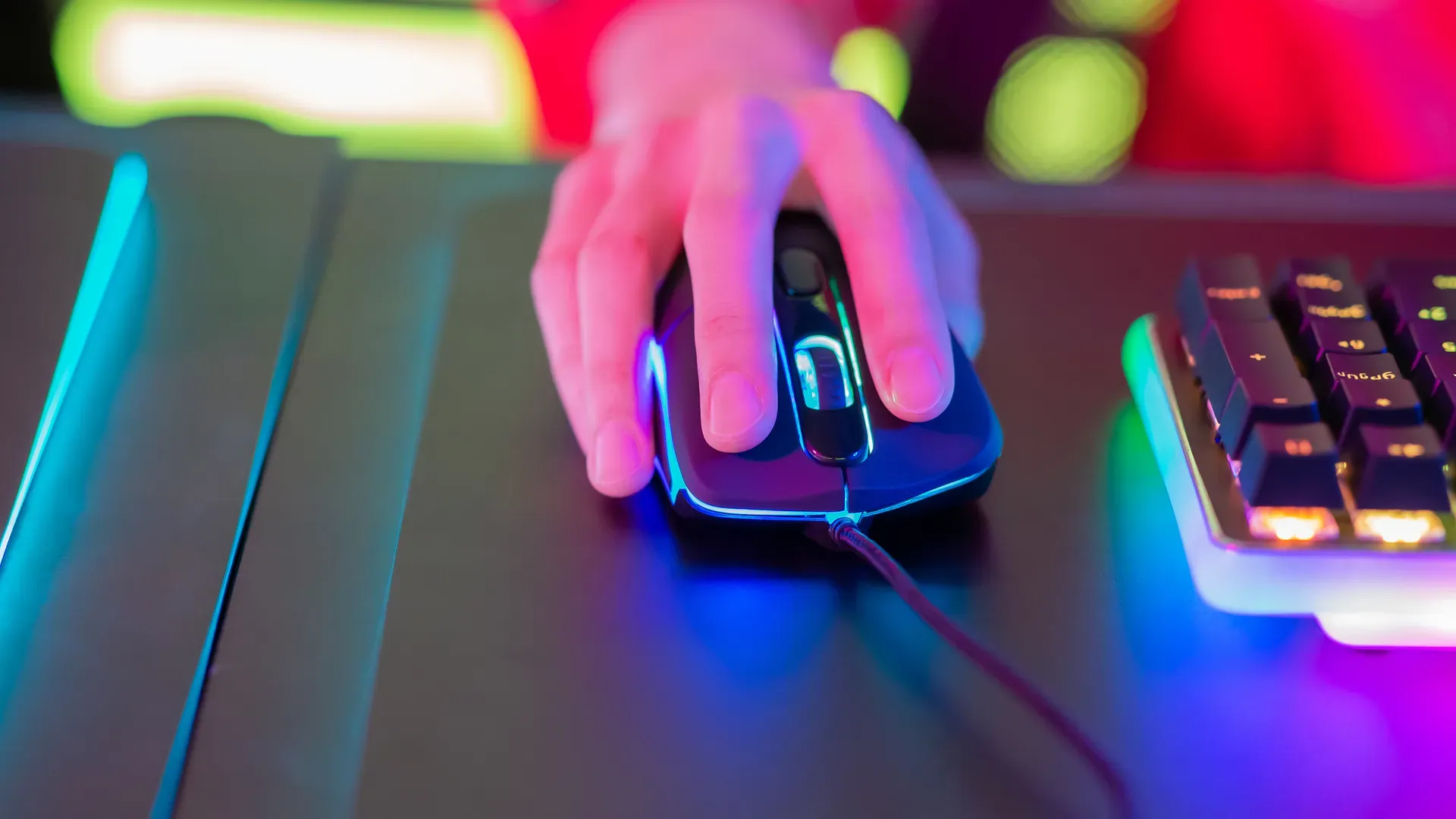 Uma mão segura um mouse gamer enquanto joga. O fundo é meio borrado e muito colorido.