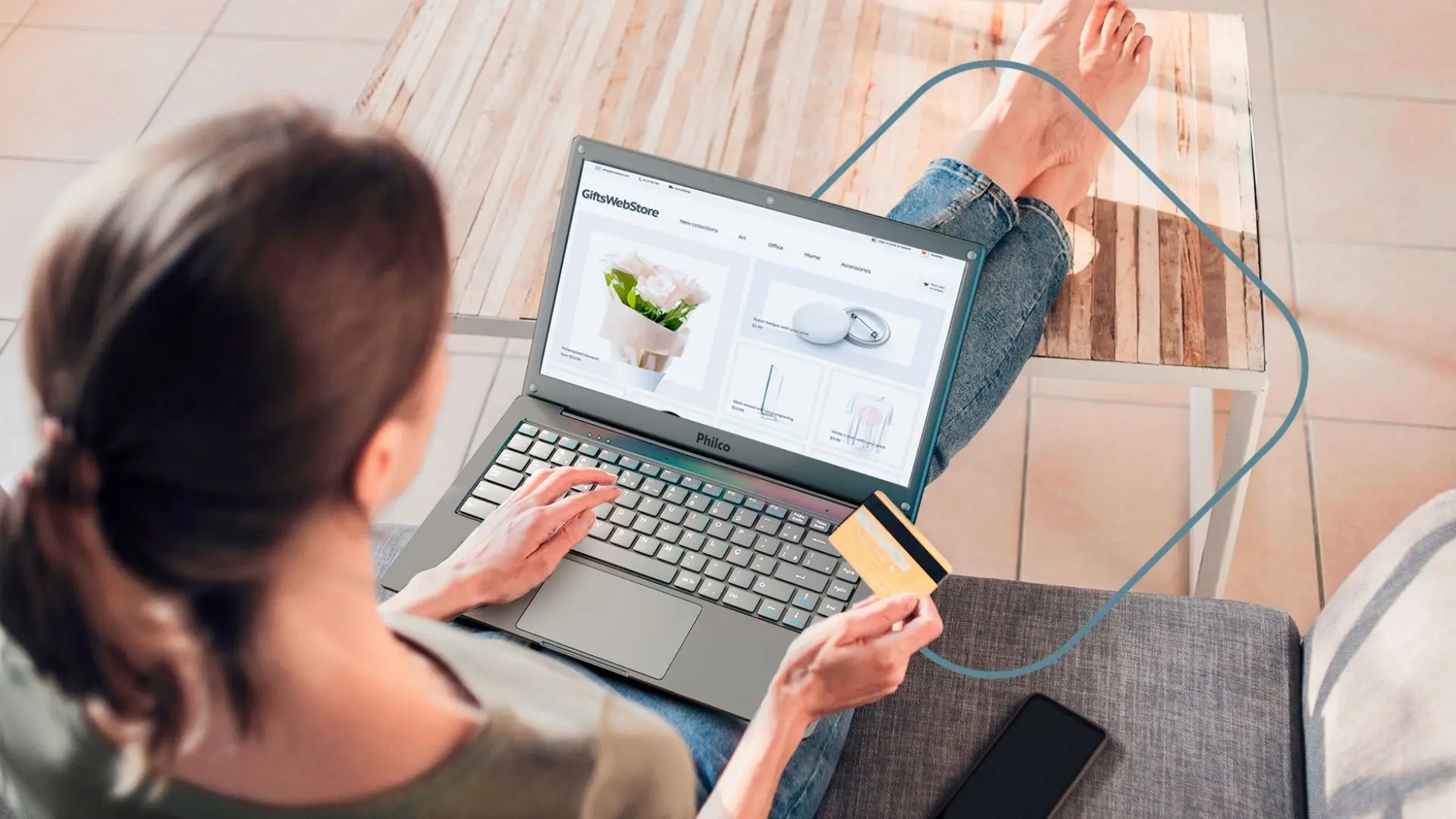 Uma mulher de calça jeans, sentada no sofá, enquanto realiza uma compra online com o notebook Philco que está em seu colo.