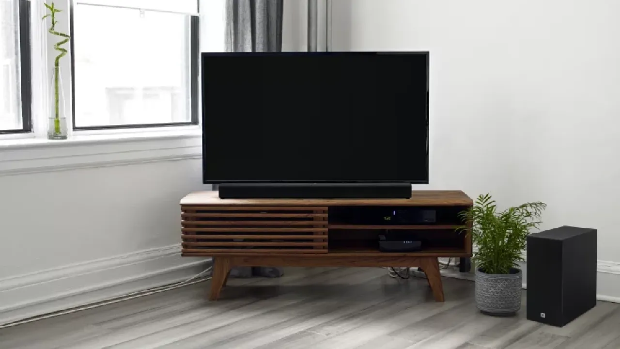 JBL SB160, uma soundbar custo-benefício, sobre rack de madeira, em frente a uma TV grande