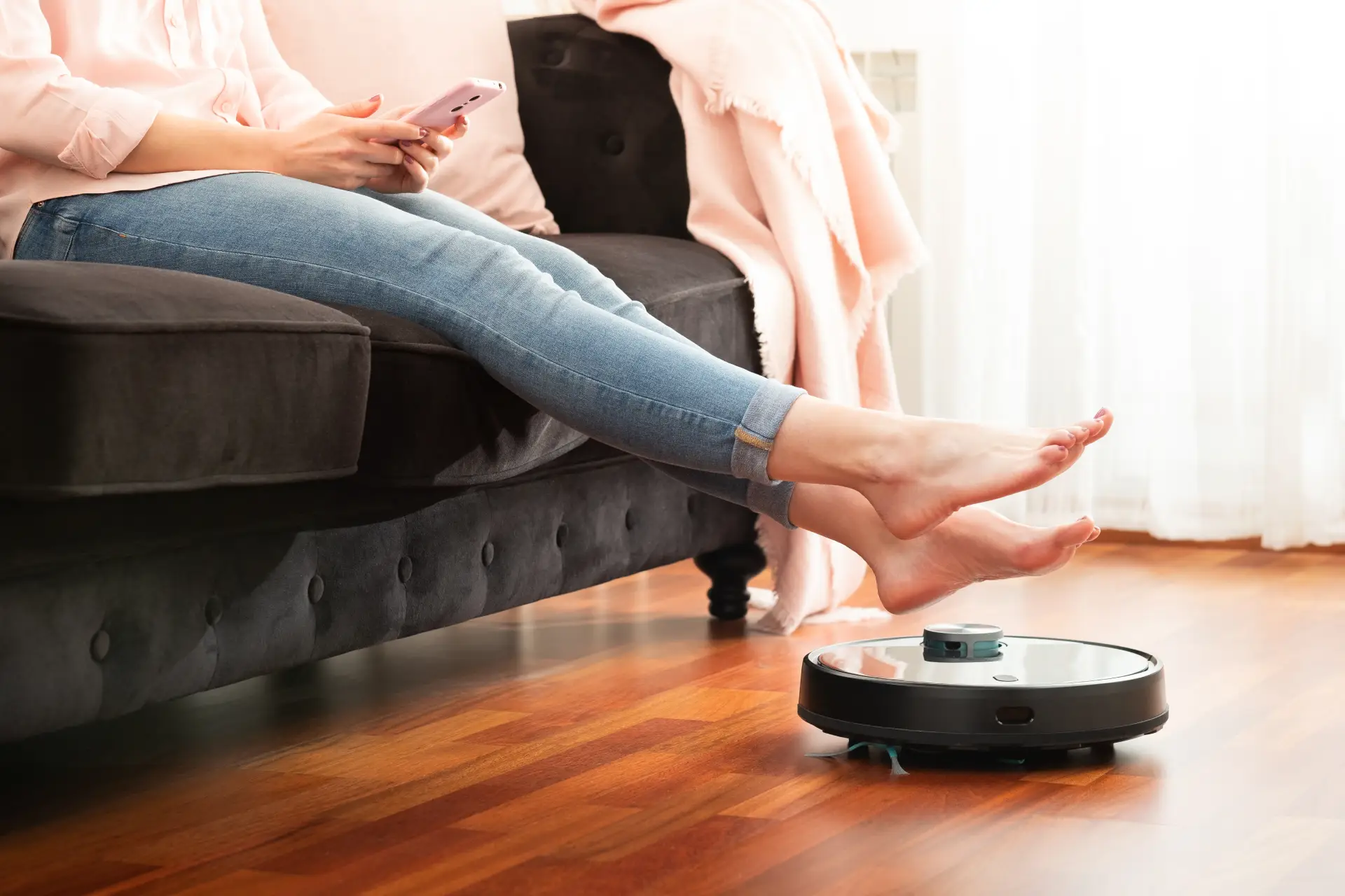 Aspirador de pó robô no chão da casa enquanto mulher sentada no sofá com as pernas esticadas controla o aparelho no celular