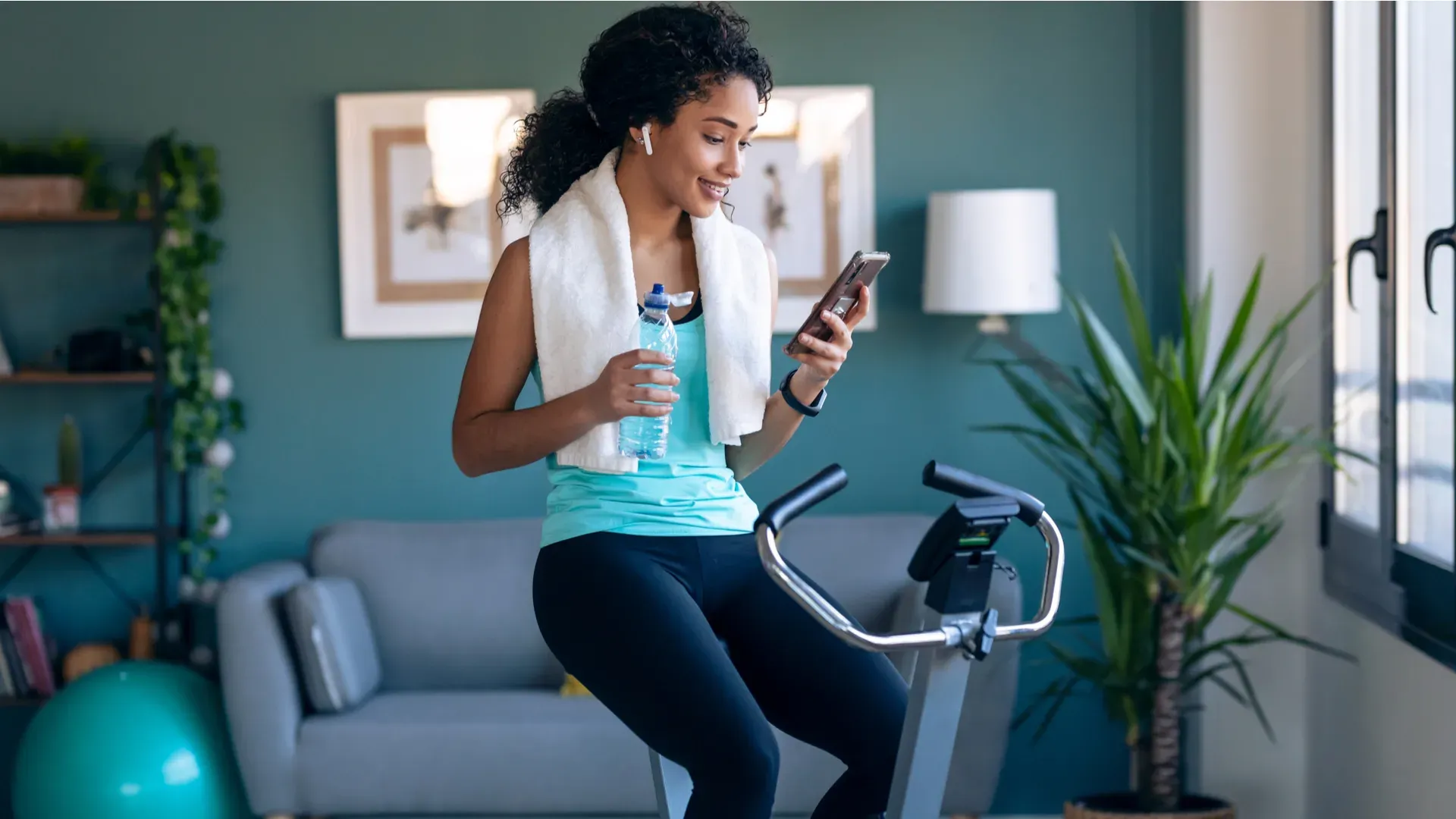 Mulher negra olhando o celular e segurando uma garrafa de água enquanto usa bicicleta ergométrica