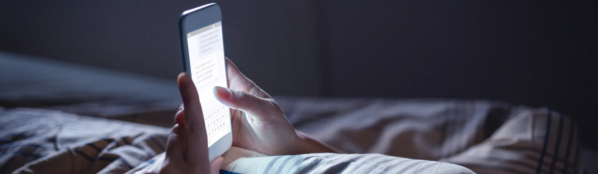 Mulher usa celular deitada na cama com luzes apagadas e apenas a tela do aparelho acesa 