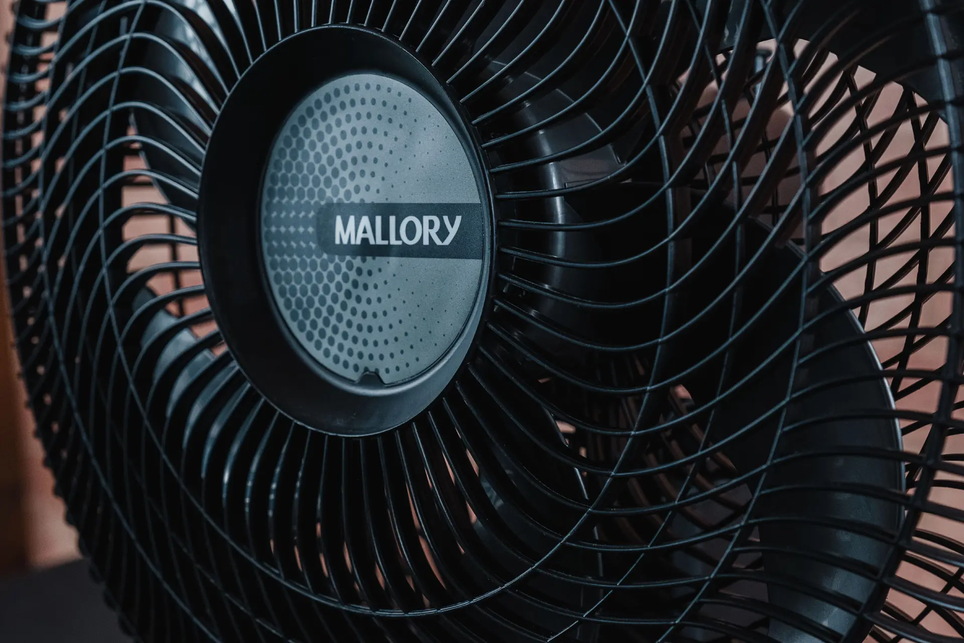 Close da grade preta do ventilador com o logo da marca no meio; a hélice do aparelho aparece ao fundo e é cinza