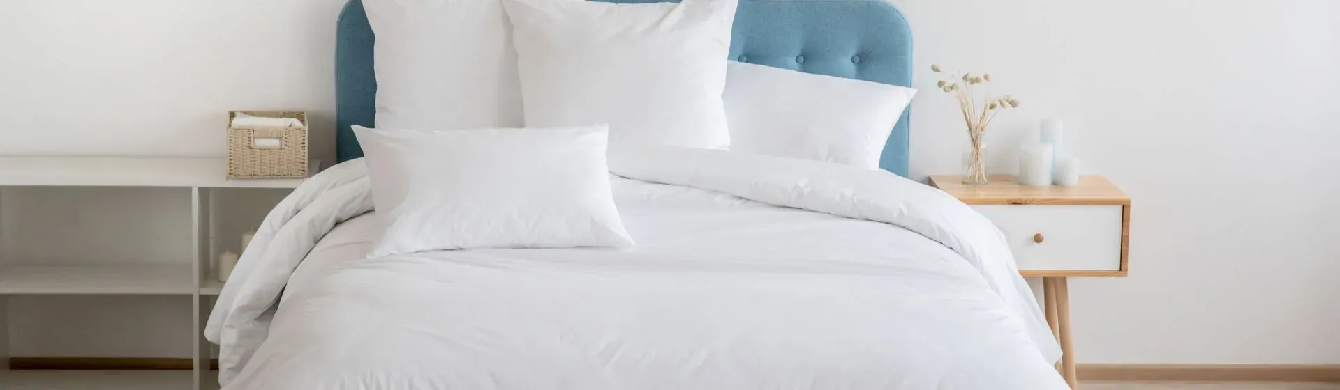 Colchão de casal com edredom e travesseiros brancos espalhados na cama, com cabeceira azul clara e móveis nas laterais