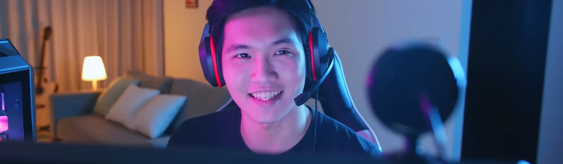 Jovem asiático jogando enquanto usa um headset gamer, com seu setup em sua frente, mas embaçado.