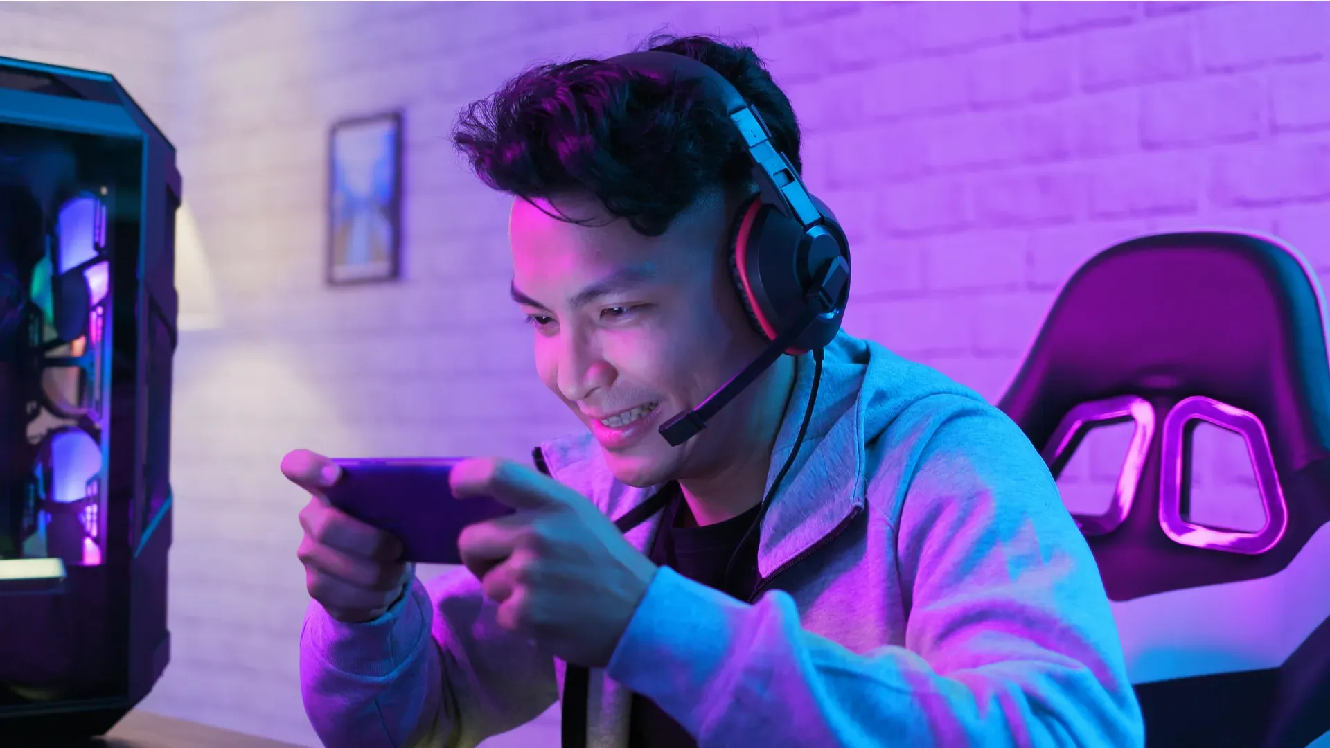 Homem asiático sentado em uma cadeira gamer joga no celular enquanto utiliza um headset, também gamer.