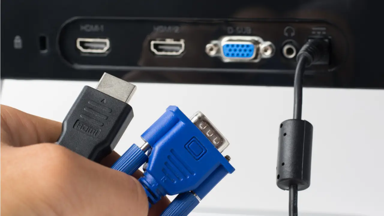 Uma mão segura um cabo HDMI e um VGA, enquanto se prepara para conectá-los ao monitor.