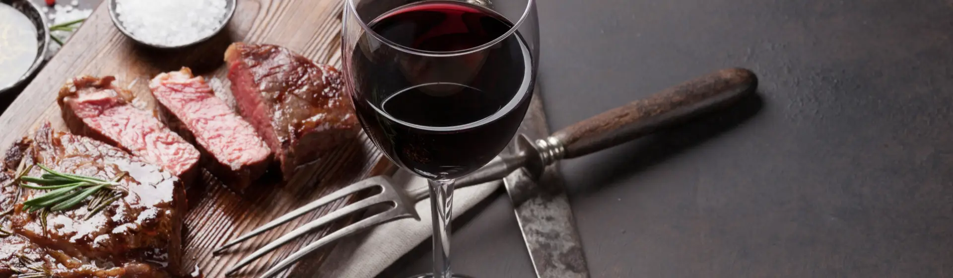  Detalhe de vinho tinto em uma taça ao lado de tábua com carne fatiada e talheres.
