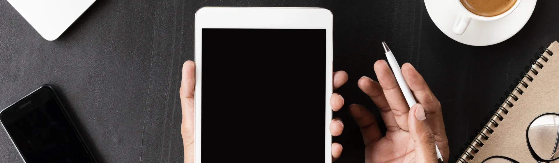 Detalhe de mão segurando iPad com tela preta, tendo a mesa de trabalho em volta