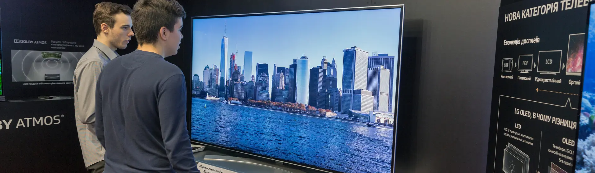 Dois homens em pé em frente a uma TV LG exibindo imagem de paisagem de cidade litorânea na tela