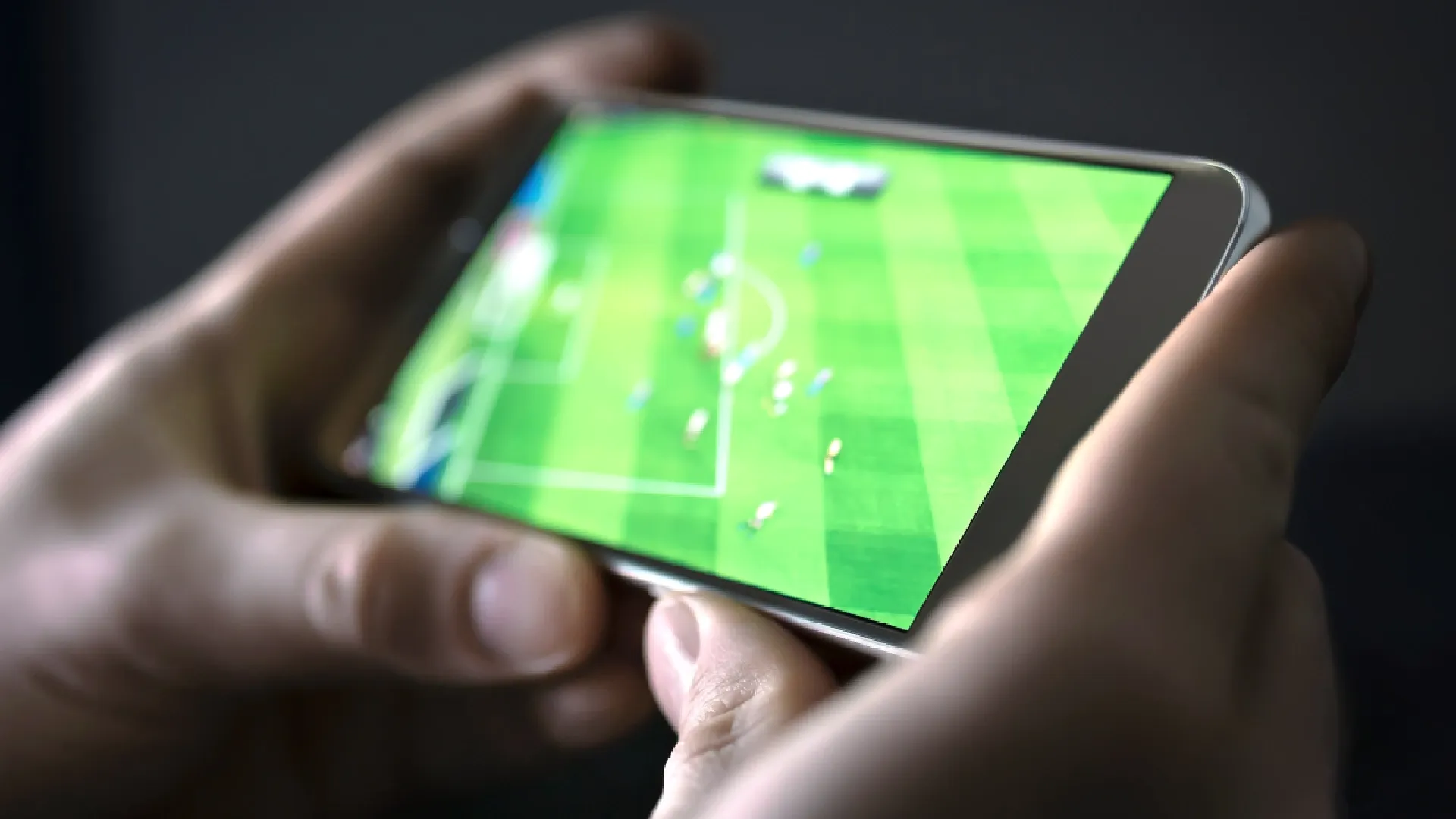 celular com tela ligada em jogo de futebol e duas mãos segurando o smartphone