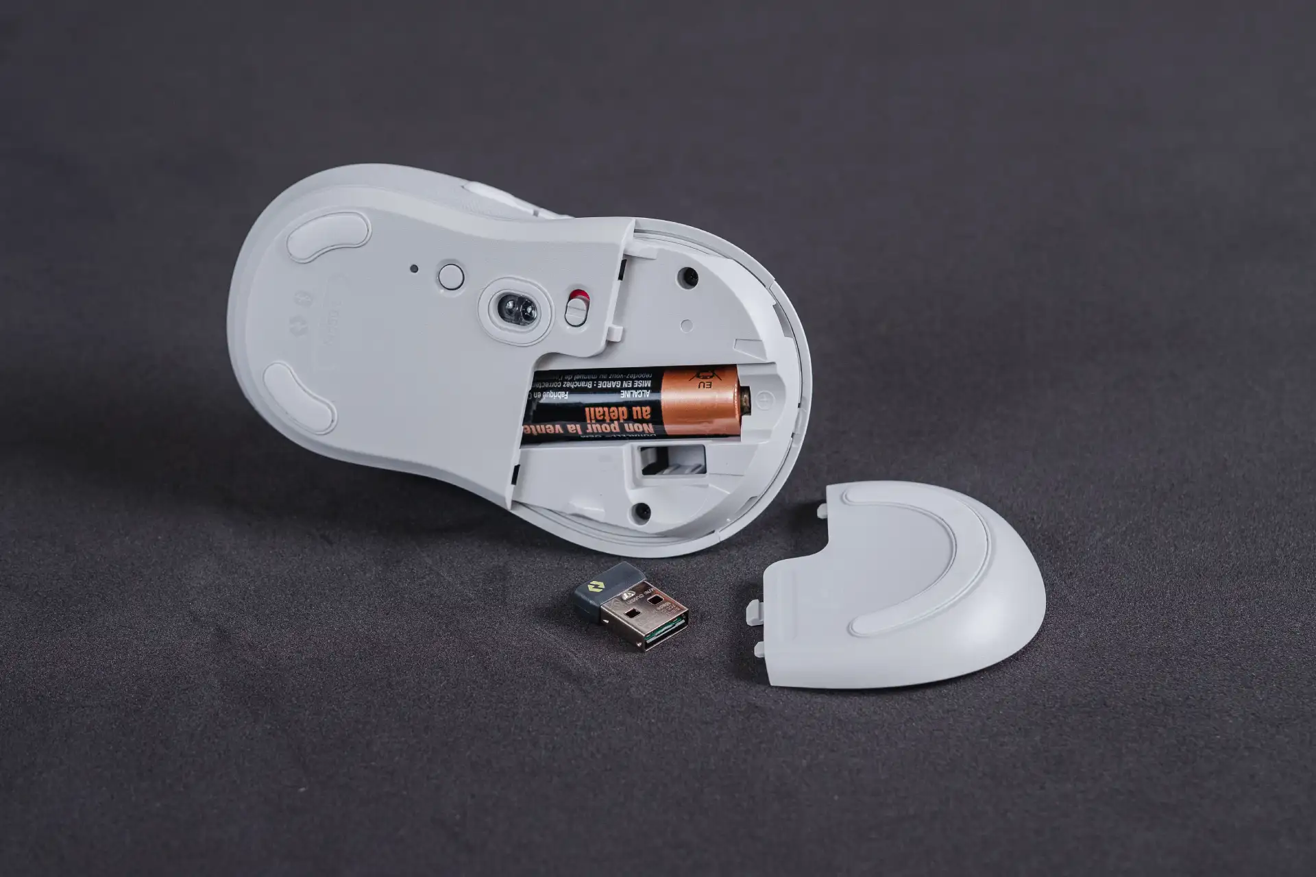 Parte inferior do mouse Logitech M650L com porta aberta, pilha e conector USB sobre mesa preta