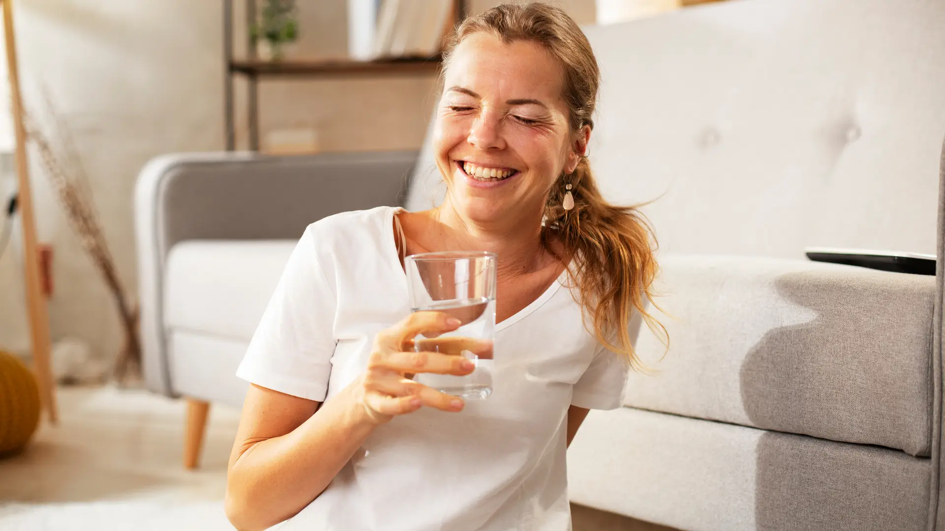 Mulher branca sentada na sala sorrindo com copo de água na mão